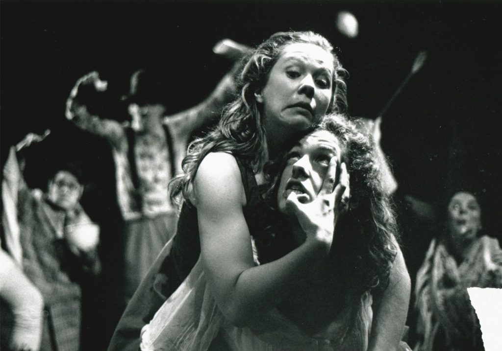 This sepia coloured image displays two women holding onto each other. The women above her is holding onto the other woman's faced. Figures are seen in the background raising their arms.