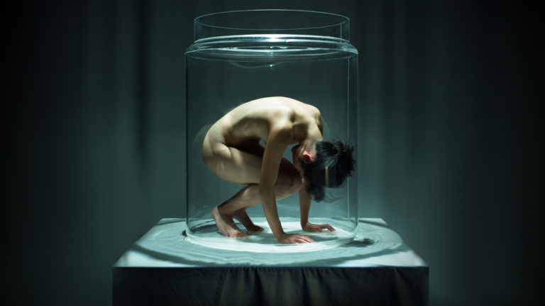 A woman placed on a table under a glass vitrine with her knees bent and arms placed flat on the table. Behind her is a dark grey curtain.