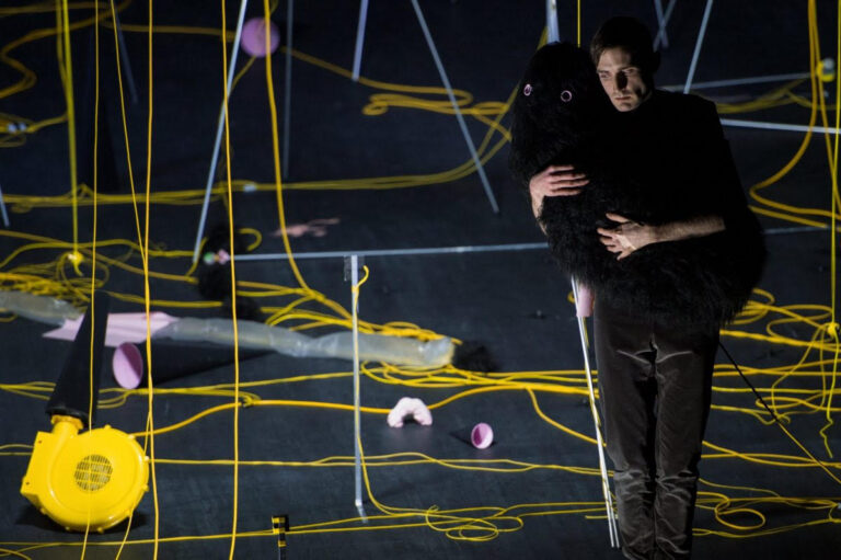 man stood on a stage hugging a fluffy black animal like object. He is surrounded by a web of yellow wires and other objects are littered around the space behind him, however we don’t know what the objects are.
