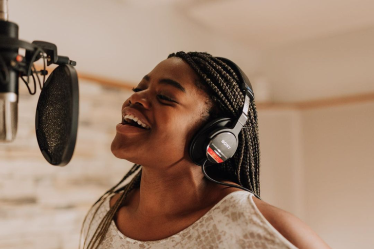This image shows the singer Tehillah Alphonso singing into a microphone in a recording studio. She is facing to the left and smiling as she sings.