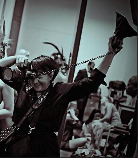 This is a black-and-white photograph of Ambra Vernuccio, a photographer. She is facing towards the camera but stood at an angle and is taking a photograph of someone else. She is holding an old-style flash bulb to the sky with her left hand and holding a camera to her right eye with her right hand. She is smiling. She is wearing a black dress with a belt at the waist, and she also has a strap over bag over her left shoulder. Behind her we see people dressed up in glamorous clothing for a burlesque show, though this image appears to have been taken backstage rather than during a performance.