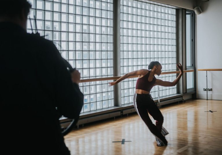 A dancer in a studio holding a pose with their arms stretched out. A figure is blurred in the shot with their back turned to the camera.