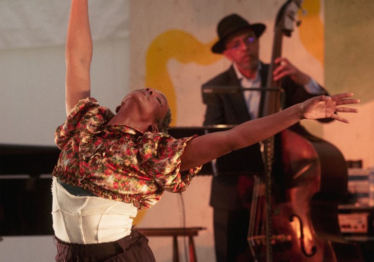 A woman and a man dancing with arms raised. There are two band members playing string instruments behind them.