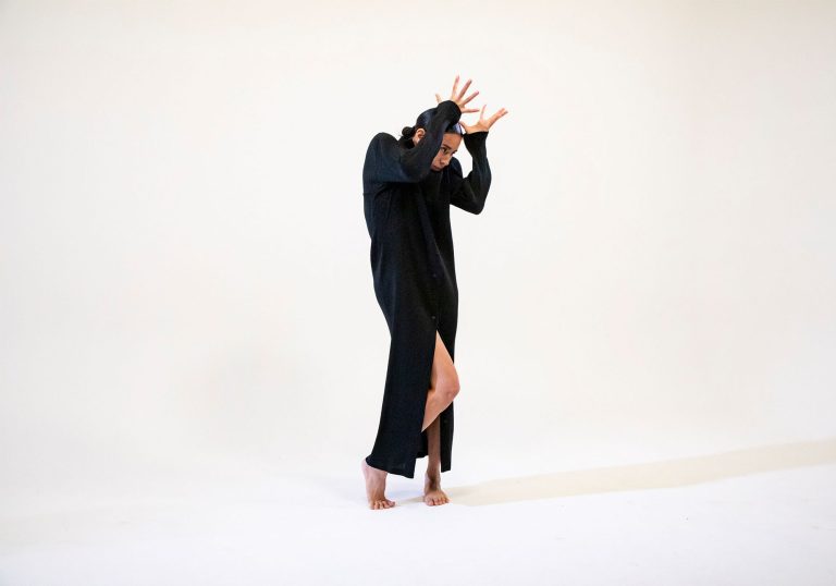 This image displays associate artist Fukiko Takase mimicking the position of a flower. Her hands are raised above her head and she is wearing an all black dress against a white background.