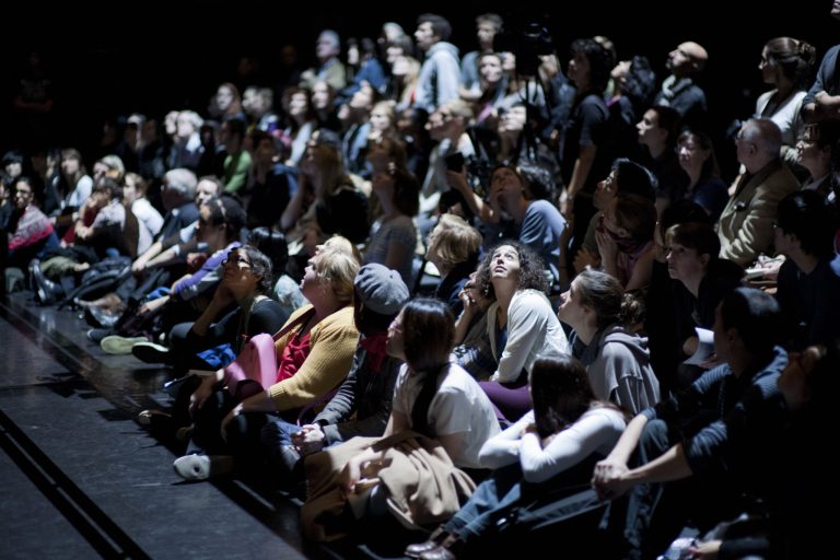 A crowd seated in a theatre auditorium, facing left