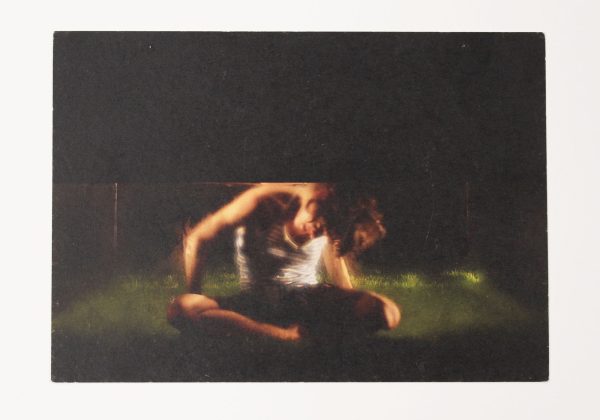 A postcard. Image features someone sat on a grassy floor, surrounded by darkness, with a low ceiling forcing them to keep their head bent downwards.